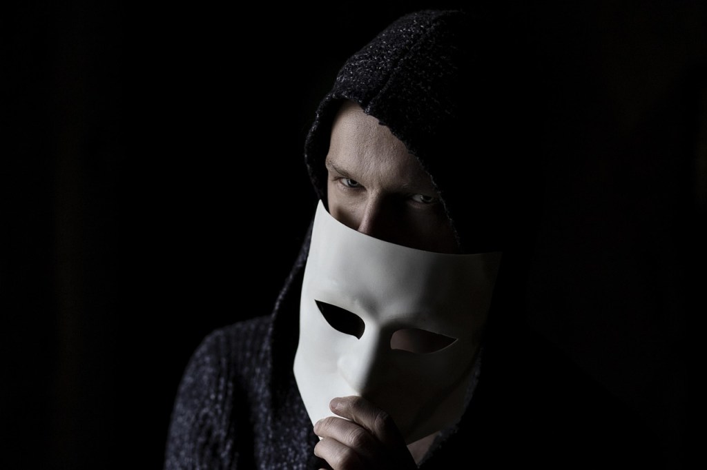 A man on a dark background removes a white mask. Only his eyes are visible.