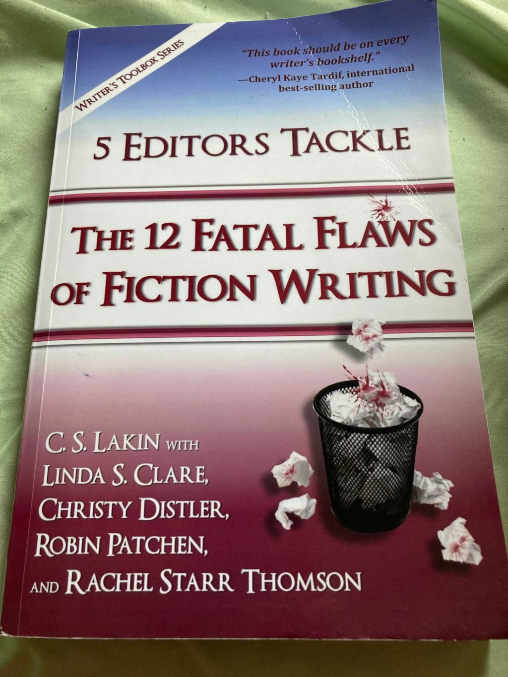 A book is seen on a light green background. The book is titled "5 Editors Tackle the 12 Fatal Flaws of Fiction Writing"