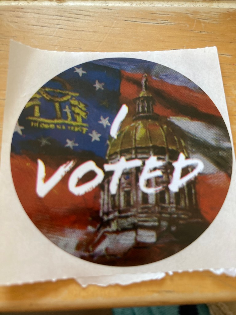An I Voted sticker with the state capitol gold dome in front of a blurry, stylized state flag. A downgrade from the peach.